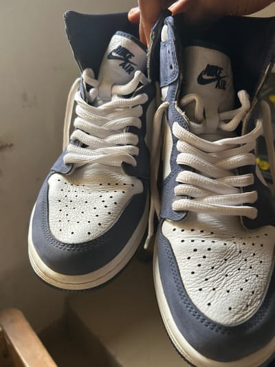 The image displays a pair of high-top sneakers, predominantly in off-white/cream and a dark blue-grey color scheme, viewed from an overhead perspective in an indoor setting. A person's fingers are visible on the left side, holding the top shoe.

The sneakers feature white laces tied in a traditional criss-cross pattern. The toe box and side panels are made of an off-white, textured material with visible perforations, suggesting leather. The overlays, mudguard, and ankle collar are crafted from a contrasting dark blue-grey material, possibly suede or a similar matte finish. The midsoles have an off-white or slightly aged cream appearance, contributing to a vintage aesthetic.

Inside the shoes, specifically on the insoles, the "NIKE AIR" logo is clearly visible in black text against a light background. The shoes appear to be in good condition, possibly being displayed or examined. The background is simple and indistinct, showing a light surface beneath the shoes and a partial view of a darker, possibly wooden, object to the right. The lighting is even, suggesting an indoor environment without specific time-of-day indicators. No specific location cues are visible within the image to denote Ashipa, Nigeria, other than the provided context.