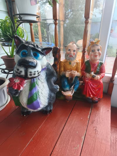 A life-sized, colorful garden gnome statue of a cartoonish wolf is positioned on a red wooden porch, holding a red and white flower and wearing a purple and green striped tie. To the right of the wolf, two more garden gnome statues are seated on what appears to be a bench. The gnome on the left is an elderly man with a grey beard, wearing a yellow shirt and blue pants, and holding a small object. The gnome on the right is a woman with blonde hair tied up with a yellow ribbon, wearing a red dress and a green vest, and holding a small, light-colored bird. Behind the statues, wooden porch railings and a glass window reveal a green outdoor space with foliage and fencing. The scene is outdoors, possibly on a residential porch in Burayevo, Russia.