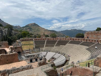 The image depicts the ancient Greco-Roman theater in Taormina, Italy, a prominent historical site situated on a hillside overlooking the Ionian Sea and Mount Etna. The theater is characterized by its semicircular seating area, constructed from stone and tiers of benches, which are partially covered by temporary white seating for modern events. In the foreground, remnants of brick walls and stone structures are visible, suggesting the ancient origins of the site. Scaffolding and temporary lighting equipment are erected in sections of the theater, indicating ongoing preparations or maintenance for performances. Several tourists are scattered throughout the seating area and walkways, observing the architecture and the panoramic views. The background features a lush, mountainous landscape with scattered vegetation and scattered buildings nestled into the hillsides, under a partly cloudy sky. The overall scene captures a blend of ancient history, modern adaptation, and natural beauty.