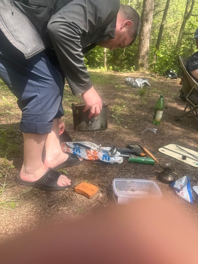The image, taken outdoors in Папярнянскі сельскі Савет, Belarus, features a man seated on the ground with his legs extended and feet wearing black slide sandals. He is holding a dark-colored camping pot in his hands. The man is dressed in a gray jacket and blue pants. Surrounding him on the ground are various camping-related items, including a green bottle, a white cutting board, a metal pitcher, a green gas canister, a small axe, a plastic bag with a logo, a plastic container with vegetation, and an orange sponge. The ground is covered in dry grass, dirt, and some sparse greenery. A portion of the image is obscured by a blurred orange object.