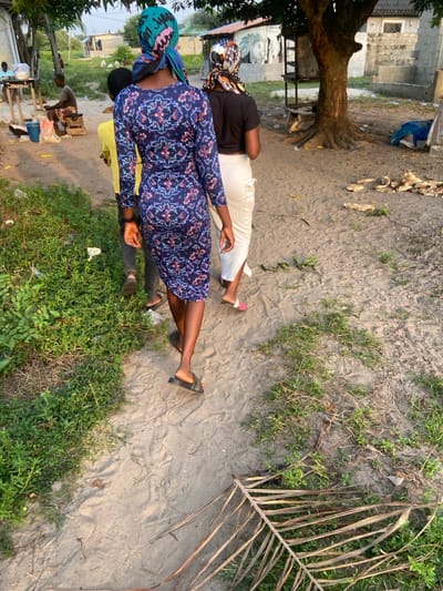 Four individuals stand on a sandy, grassy area. Two women are prominently featured: one in a navy blue floral dress and head wrap, and another in a black shirt and white pants, also with a head wrap. Two other people in a yellow shirt and light-colored shirt are partially visible. A large tree with exposed roots stands nearby, with a building faintly visible in the background. Debris, including husks, litters the sandy ground. A dried palm frond rests on the sand.