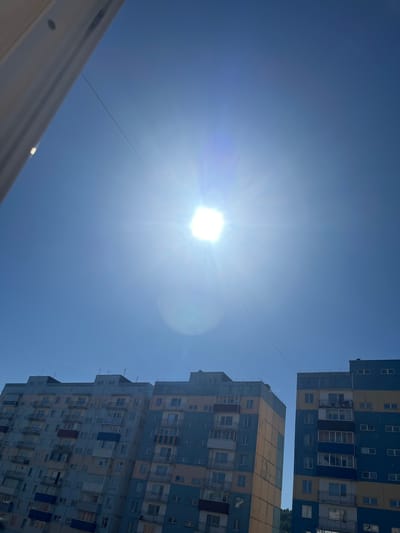 The image shows a bright, sunny day in Sheregesh, Russia, with a clear blue sky dominating the upper portion of the frame. The sun is intensely bright, creating lens flare and a halo effect. A thin wire stretches across the sky, bisecting the sun. In the lower part of the image, two multi-story apartment buildings are visible. The building on the left has facades painted in shades of blue and yellow-orange, with numerous balconies. The building on the right, also featuring blue and yellow-orange sections, appears slightly closer and taller. The architecture suggests a residential area. The scene is devoid of any people or specific activities. The environment is urban, characterized by the apartment buildings against the backdrop of the bright sky.