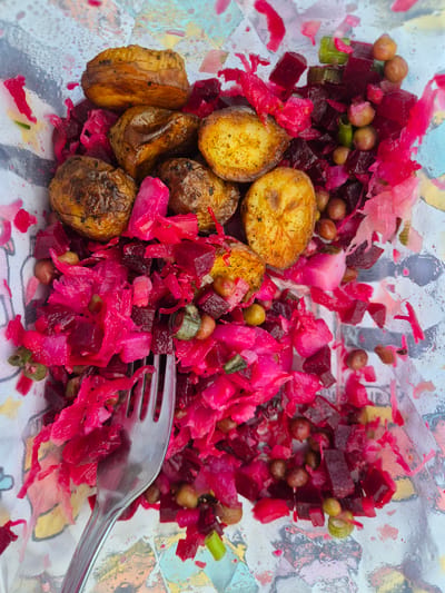 A close-up, top-down shot captures a dish of salad in a clear container, with a fork resting on the right side of the container. The salad is a vibrant mix of chopped beets, shredded red cabbage, chickpeas, and small, golden-brown roasted potatoes. The beets and cabbage lend a strong pink and deep red hue to the dish, while the chickpeas provide a speckled brown contrast, and the potatoes offer a warm, earthy tone. The salad appears to be served on a surface with a colorful, abstract pattern, possibly a placemat or the container itself. The lighting suggests it is daytime, and the overall impression is one of a rustic, home-cooked meal, perhaps eaten outdoors given the colorful background. There is no visible text in the image.