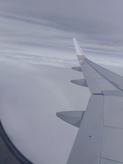 An aerial view from an airplane window shows a wing section extending diagonally across the frame. The wing is primarily white and gray, with some black lines and markings. Below the wing, a thick layer of clouds is visible, obscuring the ground. The sky above is overcast with a hazy, pale blue and gray gradient, with faint horizontal cloud formations. The scene is devoid of any people or specific landmarks that could identify a location in Hội An, Vietnam.