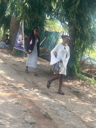 The scene depicts an outdoor location in Ijanikin, Nigeria, characterized by a dirt road lined with trees and foliage. There are two women in the photo. One wears a white dress with flared sleeves and sandals, holding a patterned bag. The other wears a grey dress with a plaid jacket and sandals, holding what appears to be a mobile phone. A banner or sign is mounted near a tree. In the background, there is a structure covered in blue tarpaulin. Another person is visible near the tree at the bottom of the frame.