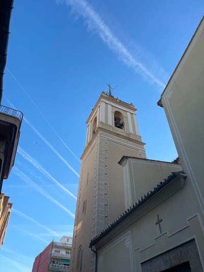 The image captures a view looking upwards in Paiporta, Spain, focusing on the town's architecture and sky. A prominent element is the bell tower of a church, showcasing its beige facade, stepped battlement-style edges, and visible bells within its arched openings. The sky is bright blue with multiple contrails from aircraft crisscrossing the expanse. Buildings flank the tower, framing the view, their facades decorated with geometric patterns, including a cross on one building. Balconies and rooftops of adjacent buildings are visible at the lower edges of the frame.