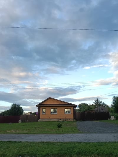 This image depicts a rural scene in Smolino, Russia, under a dramatic cloudy sky. The primary subject is a simple, tan-colored wooden house with a dark brown roof and trim. Two windows are visible on the front of the house, reflecting the sky and surrounding environment. The house is set back from a gravel driveway and a grassy lawn.

To the left of the house, a dark brown wooden fence runs along the property line, partially obscured by trees. To the right of the house, a darker, weathered wooden fence with a gate encloses a yard area, behind which a dilapidated wooden structure, possibly a shed or barn, is visible. More trees and foliage line the background, suggesting a wooded or rural setting.

The time of day appears to be late afternoon or early evening, as indicated by the soft, diffused light and the presence of clouds. The weather is overcast, with a mix of gray and lighter, more blue-tinted clouds, creating a somewhat subdued atmosphere. There is no visible activity or event taking place; the scene is static and peaceful.

Notable details include the power lines crisscrossing the sky, a common sight in rural areas. The overall impression is one of quiet, rural life in Russia. No text is visible in the image.