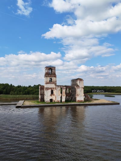 This is an aerial, vertical, eye-level, full shot of the ruins of the Church of the Nativity of the Virgin Mary in Krokhino, Russia. The church, built of red brick and white plaster, is partially submerged in the Sheksna River. The structure features a tall bell tower and arched windows. Scaffolding is present around a section of the ruins, indicating ongoing preservation efforts. A wooden pier extends from the shore to the island where the church stands. Across the water, the word "КРОХИНО" is spelled out in large, white, three-dimensional letters, serving as a landmark. The background consists of a dense green forest and a body of water that reflects the sky. The sky above is a bright blue with scattered white cumulus clouds, and a faint contrail from an airplane is visible. The overall scene depicts a historic landmark in a state of picturesque decay, situated within a natural landscape.