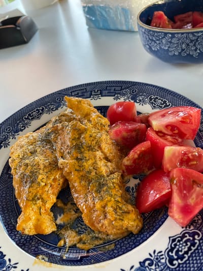 This is a top-down, close-up photograph of a meal served on a plate. The main subjects are two pieces of cooked fish, coated in a creamy, orange sauce and sprinkled with chopped green herbs, likely dill. The fish is presented on a blue and white patterned plate, in the style of Willow pattern. To the left of the fish, on the same plate, are several wedges of ripe, red tomatoes, some of which appear to be cut from cherry tomatoes.
In the background, to the left, is a blue and white patterned bowl containing more tomato wedges. The setting appears to be a table, with a white surface visible to the left of the plate. A partially visible black object, possibly a small appliance or container, is also in the lower left corner of the frame.
The lighting suggests it is daytime. There are no people visible in the image. The meal appears to be freshly prepared and ready to be eaten. The overall impression is of a simple, homemade meal. No visible text is present. The location context is Skala Avdiron, Greece.