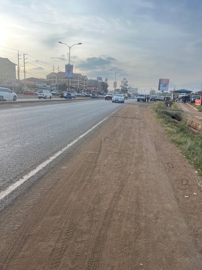 A photograph shows a slightly tilted view of a road with moderate traffic. The road is damp and shiny, with a white line dividing it from a dirt shoulder. Buildings and billboards line the left side of the road. Tire tracks are visible on the dirt shoulder. Overhead power lines stretch toward the sky. The sky is cloudy with hints of blue.