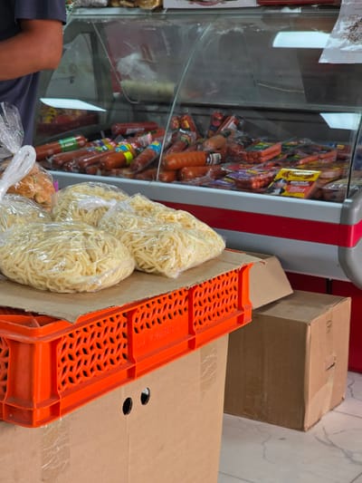 In an indoor setting, likely a small store or market in Talgar, Kazakhstan, various food items and boxes are visible. In the foreground, several packages of dried noodles are prominently displayed, wrapped in clear plastic. To the left, a glass display case is filled with a variety of sausages and other cured meats. The sausages are packaged and hang from racks, some with colorful labels. A person's arm and knee are visible in the lower left corner, suggesting their presence in the scene, though their face is not shown.
Behind the noodle packages and to the right, a bright orange plastic crate is placed on top of cardboard boxes. The crate has a distinctive lattice pattern. More cardboard boxes are stacked in the background. The overall scene appears to be a retail environment where food products are being stored or displayed. The lighting suggests it is daytime. No specific weather conditions are discernible. There is text visible on some of the sausage packaging, but it is not legible enough to be identified. The phrase "AO TEHb ANPO" is visible on the orange crate, printed in black text.