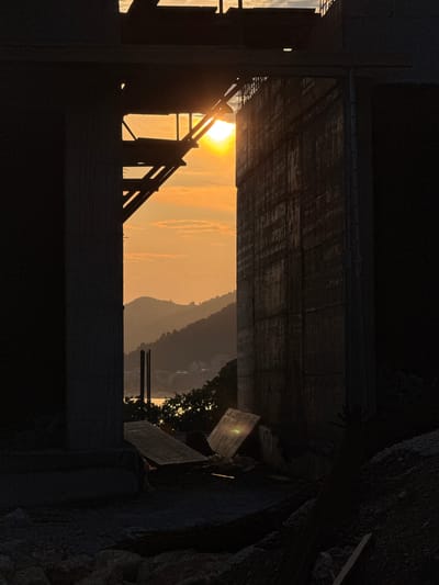 The image captures a sunset viewed through an opening in a construction site, framed by large concrete structures. The sky is a vibrant orange and yellow, with scattered clouds. The sun is partially obscured by scaffolding and appears as a bright orb. In the distance, silhouetted hills descend towards the sea, with faint lights visible on the coastline, suggesting the presence of a settlement. The foreground is dominated by dark, rough-textured concrete pillars and walls, characteristic of unfinished construction. Construction debris, including wooden planks and what appears to be rebar, litters the ground. The overall scene conveys a sense of raw, in-progress development against the backdrop of a serene natural landscape and the fading light of day. The location is identified as Vrijesno, Montenegro. There are no people visible in the image.