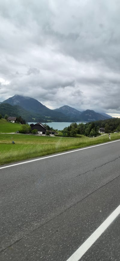 The image shows a scenic view of Hof bei Salzburg, Austria, with a body of water, mountains, and rural buildings. The foreground features a gray asphalt road with white lane markings. Beyond the road, a lush green field slopes upwards, dotted with trees and a few houses. In the middle ground, a large, dark-colored building with a gabled roof is prominent. Further back, a turquoise lake is visible, surrounded by dense green forests. Majestic mountains, partially obscured by dark, dramatic clouds, form the backdrop. The sky is overcast, suggesting a cloudy or potentially rainy day. A person is visible riding a bicycle in the field. The overall impression is one of natural beauty and tranquility.