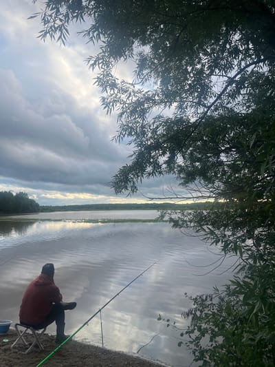 A person is fishing on the bank of a body of water, with trees and foliage overhead. The person is seated on a small folding chair, facing the water, and is wearing an orange jacket and dark pants. Two fishing rods are visible; one is in use by the person, and another is set up further down the bank. The water is calm and reflects the sky, which is partly cloudy with some patches of sunlight. The time of day appears to be late afternoon or early evening, with the sun low on the horizon. The weather is overcast but not raining. The scene suggests a peaceful and solitary fishing activity. No text is visible in the image.