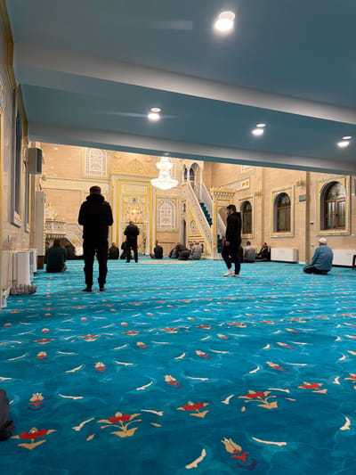This image depicts the interior of a mosque located in Breznitsa, Bulgaria. The scene shows a large prayer hall with a dominant blue carpet featuring stylized red and yellow floral patterns and white accents. The walls are light tan/cream, elaborately decorated with golden trim and intricate white carved designs, particularly around the mihrab area at the far end. A large, multi-tiered crystal chandelier hangs from the ceiling in the background. To the right of the mihrab, a minbar (pulpit) with green carpeted stairs and golden railings is visible. Recessed lighting illuminates the light blue and white ceiling. Arched windows with dark wood frames are visible on the right wall.
Approximately ten to twelve adult males are present in the prayer hall. In the foreground, one male dressed in a dark jacket and trousers stands with his back to the viewer, facing the mihrab. Another male, similarly dressed in a dark jacket and trousers, walks from right to left in the middle ground, wearing white-soled shoes. Several other males are seated on the carpet in various positions, primarily facing towards the mihrab; some are kneeling or sitting with their legs tucked, while one near the right wall wears a light blue shirt and a white cap. A white rectangular wall unit is visible on the left wall, and similar units are along the right wall. A digital clock is mounted on the right wall. The overall environment suggests a space for worship or communal gathering.