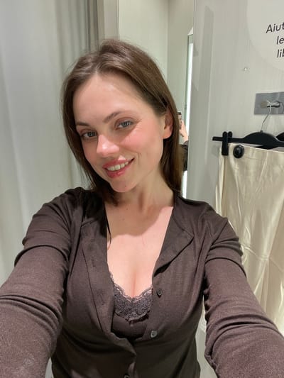 A female subject is depicted in a contemporary fitting room located in the city of Pompei, Italy. The subject, appearing to be in her 20s or 30s with long brown hair, is shown from the chest up, smiling directly at the camera, which is reflected in a mirror to her left, indicating a self-portrait. She wears a dark brown long-sleeved button-down cardigan over a dark brown, lace-trimmed camisole. The environment consists of light-colored walls, with a large mirror occupying the left side of the frame and a clothing rack with a black hanger holding a light-colored garment (possibly pants or a skirt) on the right. Partially visible Italian text is present on the upper right wall.