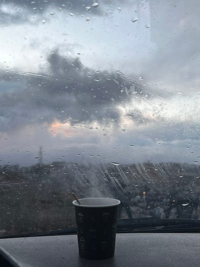 The image depicts an interior view from a vehicle looking out through a rain-streaked windshield, set in Dummar Municipality, Syria. In the foreground, a dark-colored disposable coffee cup with white outlines of coffee cups and beans rests on a grey dashboard. Steam emanates from the cup, which contains a light brown stir stick. The windshield is covered extensively with numerous raindrops and vertical streaks. Through the wet glass, a cloudy sky dominates the upper background, showing hints of orange and pink light near the horizon, suggesting either dawn or dusk. The lower background reveals a blurred landscape, possibly a distant urban or natural environment, with faint, scattered lights visible. No individuals are directly visible.
