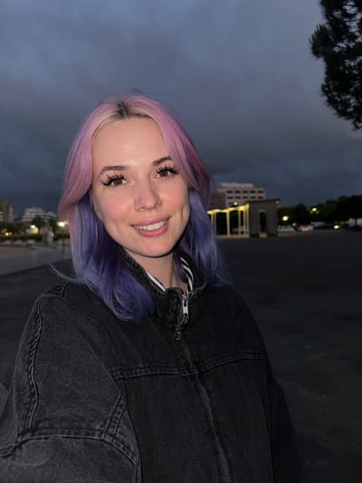 A cheerful young woman is captured in a close-up, waist-up portrait, smiling broadly at the camera. She has striking multi-toned hair, primarily shades of pink and purple, with some darker roots visible. Her eyes are open, revealing light brown irises framed by prominent, long false eyelashes and well-defined eyebrows. She wears a dark, possibly black or dark grey, denim jacket with visible seams and a metallic zipper detail on the front, over what appears to be a striped garment underneath. Her skin appears fair and smooth, with a subtle blush on her cheeks.
The setting is an urban environment at dusk or night in Gijón, Spain. The sky in the background is a deep, dark blue-grey, indicating low light conditions. Behind the woman, several multi-story buildings are visible, illuminated by scattered artificial lights that appear as soft glows. Trees, silhouetted against the dark sky, are also present in the upper left portion of the image. The overall atmosphere is dim but punctuated by the warm glow of city lights. No text is visible in the image.