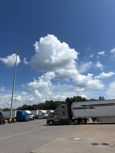 A sunny daytime, low-angle exterior shot captures a parking lot filled with commercial trucks and trailers under a bright blue sky dotted with large, white cumulus clouds. A prominent utility pole with multiple wires stands to the left of the frame. Several semi-trucks, some blue, some white, and a dark grey one in the foreground, are parked in rows. The grey truck, a Freightliner model, is equipped with a white trailer featuring a distinctive black and orange logo and stripe design. The parking lot surface is asphalt with yellow line markings. In the background, a line of green trees forms a treeline. The scene is consistent with a truck stop or a depot.
