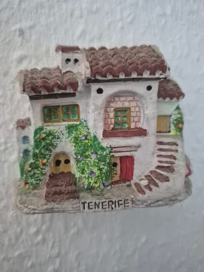 The image is a close-up, eye-level photograph of a small, decorative model of a house, likely a souvenir. The house is white with a reddish-brown tiled roof. It features multiple windows, some with green panes and brown frames, and a red door. Green vines with small purple flowers and red and yellow accents climb the side of the house, near a brown, arched doorway. A stone staircase, also in reddish-brown tones, leads up to a doorway on the right side of the house. The word "TENERIFE" is printed in black capital letters on the base of the model. The background is a textured, off-white surface, suggesting the model is attached to a wall or other surface. There is no indication of the city of Trier, Germany, in the image.