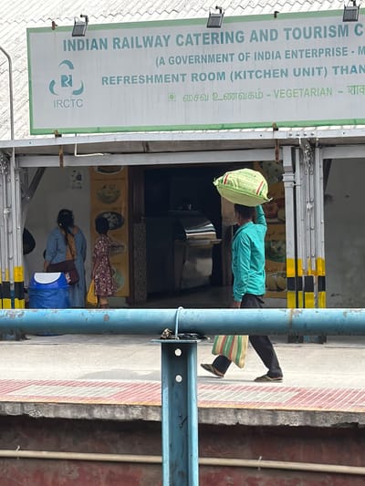 The image captures a scene at what appears to be a train station in Thanjavur, India. A prominent sign displays "INDIAN RAILWAY CATERING AND TOURISM C" and "IRCTC (A GOVERNMENT OF INDIA ENTERPRISE - M REFRESHMENT ROOM (KITCHEN UNIT) THAN VEGETARIAN - शाक".
In the foreground, a person in a teal-colored shirt and dark pants is lying down, possibly resting or sleeping, with a light green bag acting as a pillow. Their legs, clad in dark pants and flip-flops, are visible, as is a colorful striped bag placed near their feet.
Further back, through an opening, two other individuals are partially visible. One person appears to be lying down, while another is seated or in a similar resting posture. The background suggests an indoor area, perhaps part of the refreshment room, with posters or artwork on the walls. The overall lighting suggests it is daytime. The presence of the IRCTC sign and the visual of people resting strongly indicate a railway station environment. The scene conveys a sense of everyday life and rest in a public transit hub.