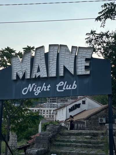 The image shows a sign for "MAINE Night Club" in Podostrog, Montenegro. The sign has large, illuminated block letters spelling "MAINE" in white, and smaller white cursive text below reading "Night Club". The sign is mounted on a dark blue background. In the background, there is a building with a sign that also reads "MAINE", with a person standing on stairs in front of it. The overall scene depicts an outdoor establishment, likely a nightlife venue, set against a backdrop of buildings and vegetation in a hilly or mountainous area.