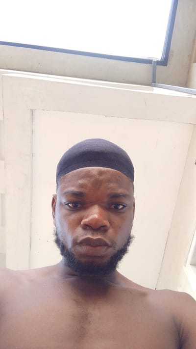 A close-up shot shows a shirtless Black man with a black cap covering his head, a short beard, and visible chest hair. He appears to be indoors, under a white ceiling with a window or skylight visible. There is no specific indication of the city of Enugwu-Ukwu, Nigeria in the image.