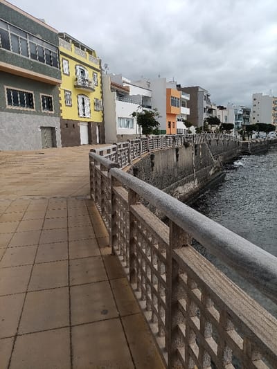 The scene depicts a coastal promenade in the city of Agüimes, Spain, under an overcast sky during daytime. The foreground features a light brown, tiled pedestrian walkway extending from the lower-left corner, curving slightly to the right. A sturdy railing separates the promenade from the water, composed of a smooth, light grey top rail and square-patterned, weathered concrete or stone infill panels.

Beyond the railing, a dark, textured seawall rises from the water, which appears dark blue-grey and choppy, with small waves breaking against the wall. The seawall shows areas of dampness and minor organic growth. Small trees or bushes are intermittently planted along the promenade side, behind the railing.

A continuous row of multi-story buildings forms the background, lining the opposite side of the promenade. From left to right: a dark grey/green building with strip windows; a prominent three-story building with a bright yellow facade, white shutters, a central balcony with an ornate white railing, and a ground-level garage door; followed by a series of predominantly white, orange, and light grey buildings featuring balconies and varied modern architectural designs. No people or discernible actions are present in the image.