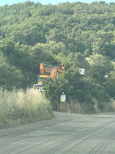 An orange excavator, labeled "JCB," is partially obscured by dense green foliage on a hillside. The excavator appears to be stationary. In front of the foliage, a dirt road curves through the foreground, with dry grass and vegetation bordering the road. A vertical marker with colored bands (yellow, green, red) stands near the road. The background is a heavily wooded hillside under a bright sky. The scene appears to be located in a rural or semi-rural area, possibly on the outskirts of Aytos, Bulgaria, given the prevalence of natural landscapes.