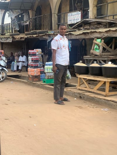 The image captures a bustling street market scene in Egbedi, Nigeria, under bright daylight. In the foreground, a dark-skinned male, approximately 25-35 years old, stands facing slightly left of the camera. He wears a white short-sleeved collared shirt with a red floral pattern, dark brown trousers, and black slide sandals. He holds a dark-colored smartphone in his left hand and a clear plastic bag containing green contents in his right hand.
To the left of the central figure, a silver motorcycle's front portion is visible. Behind it, a commercial display features tall stacks of bottled beverages, including red-labeled soft drinks in yellow plastic crates and blue-labeled water bottles in green plastic crates. Further left and behind these products, three individuals are seated on a raised concrete ledge: one male in a white traditional garment holds a small object, flanked by another male to his left, and a partially obscured figure further left, possibly wearing a head covering.
To the right of the central figure, a wooden market stall displays three large black tubs, each filled with a light-colored, granular substance. Behind these stalls, the street is lined with weathered, multi-story buildings featuring arched windows and balconies, indicative of commercial establishments. A white sign with "OSSAD COMMUNICATION" is visible on one of these buildings. Makeshift roof structures, constructed from wood and corrugated metal, are present, with several stacked tires observed on one lower roof. The ground surface is unpaved, composed of reddish-brown dirt or sand, with a concrete drainage gutter running parallel to the storefronts. A small white object lies on the ground near the gutter. No direct interactions or specific actions between the individuals are discernible.