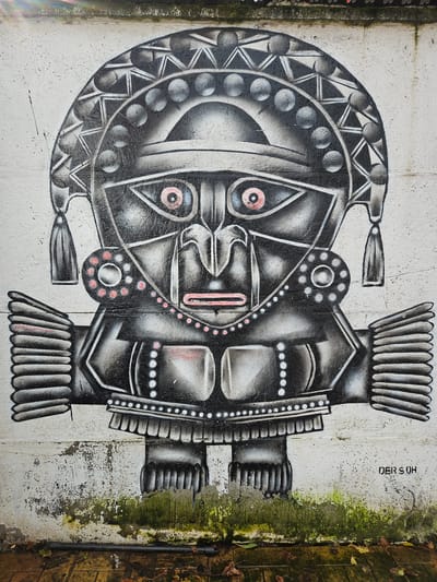 A horizontally oriented eye-level shot depicts a piece of graffiti art resembling a stylized mask, possibly inspired by indigenous or tribal designs. The artwork is painted on a concrete wall, which shows signs of wear and weathering. Moss is visible on the wall's edges, suggesting damp conditions. To the far right, a portion of a brick wall can be seen.
The mask is rendered in shades of black, white, and gray, with small touches of red. The left side of the mask features a rounded shape with geometric patterns and circular motifs. The right side is more angular and symmetrical, with a series of rectangular forms and vertical lines.
In the top right corner of the image, a signature or tag is visible: "DER S OH." The scene appears to be outdoors, possibly in an urban or semi-urban setting. The location is in the city of Osorno, Chile.