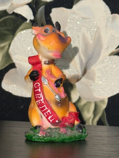 A small, orange and pink figurine of a deer-like creature stands on a green base adorned with pink flowers. The figurine has black hooves and ears, blue eyes, and a red ribbon draped around its neck and torso. The ribbon is inscribed with white Cyrillic lettering that appears to read "СТРЕЛЕЦ" (Strelets), meaning "Archer" in Russian. A silver arrow is depicted on the figurine's chest, pointing downwards. The background consists of a blurry floral pattern with white and green elements, possibly wallpaper. The figurine is placed on a dark, reflective surface, which is likely a table or shelf. The lighting suggests an indoor setting. There is no identifiable location in the city of Bender, Moldova, depicted in this image.