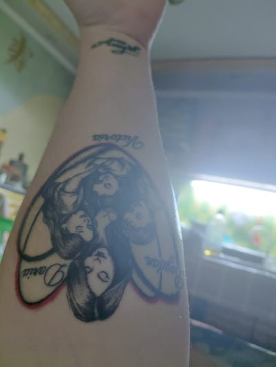 The image shows a tattoo on a person's forearm. The tattoo depicts four faces within a heart-shaped outline. The names "Victoria," "Daria," and "Bogdan" are visible, written in script below the faces. The faces appear to be rendered in black ink with shading. There is also a smaller, faded tattoo of what appears to be a bird or wing on the upper part of the forearm. The background is out of focus, showing a blurry interior environment with what looks like sunlight or artificial light coming from a window or light source. The scene does not provide any information about the location in Marynivka, Ukraine.