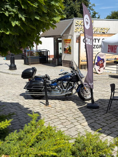 A blue and chrome cruiser motorcycle is parked on a cobblestone street in front of a building with a sign that reads "SKLEP SPOŻYWCZY" (Grocery Store). To the right of the motorcycle is a tall, dark flag with a white heart logo. There is also a white tent with red lettering and a circular advertisement featuring a cartoon chocolate bar character wearing a wizard hat. Green bushes are in the foreground, and a large tree with green leaves partially covers the upper left portion of the image. The scene appears to be set in a town square or pedestrian area in Świeradów-Zdrój, Poland, on a sunny day.