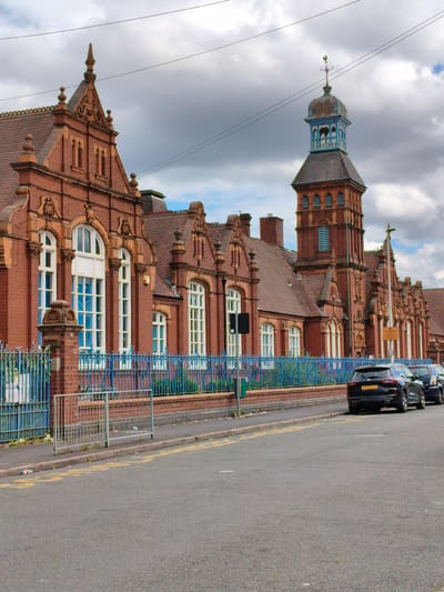 The image depicts a red brick building with a distinctive tower and a weather vane, likely a school or public building, situated in Sandwell, United Kingdom. The building features ornate architectural details, including decorative carvings and arched windows with white frames. A bright blue metal fence runs along the front of the property, separating it from a paved road.
Parked along the roadside are several cars, including a black SUV with the license plate "BG74 LLC". The street is lined with yellow markings, and a few traffic signs are visible. The weather appears to be overcast with a dramatic sky filled with clouds, suggesting it is daytime. The scene is quiet and appears to be unoccupied by people. The overall atmosphere is one of a typical urban streetscape on a cloudy day.
