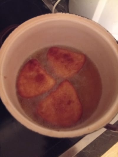 A light-colored cooking pot, approximately 30 cm in diameter, is depicted from an overhead perspective, containing hot cooking oil and three irregularly shaped, deep-fried food items in Domsühl, Germany. Each food item possesses a golden-brown to reddish-brown exterior, indicative of a frying process, and measures approximately 10-12 cm across. The oil within the pot exhibits active bubbling. The pot rests upon a dark, flat surface, likely a stovetop. Portions of a lighter-toned background, consistent with a kitchen wall or appliance, are visible. No human subjects or additional objects are identifiable.