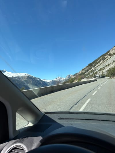 The image, taken from inside a vehicle, depicts a road alongside a steep, mountainous landscape. The road is paved, with white lane markings. The mountains are rugged, with exposed rock and some vegetation. A stretch of snowy peaks is visible in the distance. The sky is clear and blue. The interior of the vehicle, including part of the dashboard, steering wheel, and air vent, is visible in the foreground. The location is likely in a mountainous area, possibly near Montgenèvre, France, considering the Alpine landscape. There are no people visible in the frame.