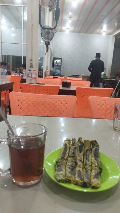 The image depicts a dining area, possibly a restaurant, in Sigli, Indonesia. In the foreground, on a table, is a glass mug filled with a dark liquid, likely tea, and a green plate containing several grilled food items wrapped in leaves. The background reveals a spacious interior with numerous empty orange plastic chairs arranged around tables. A ceiling fan is visible, along with strip lighting on the ceiling. Further back, a person wearing a black shirt and a peci hat is standing and facing away from the camera. The environment appears to be indoors, with large windows or glass panels on the left side. The overall atmosphere suggests a casual dining setting.