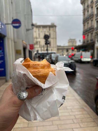 The image depicts a first-person perspective shot on a street in Vincennes, France, under an overcast sky. In the foreground, a left hand holds a partially consumed golden-brown croissant, wrapped in crinkled white paper, from which a bite has been taken from the left side. The hand wears a large silver ring on the ring finger, featuring a circular, detailed relief pattern. Partially visible text on the white paper suggests "BOULANGERIE".
The background is out of focus, showing an urban street scene. The asphalt road surface appears wet, indicating recent precipitation. Various vehicles, including passenger cars and a white utility van, are present on the road, with several red tail lights illuminated. To the left, a building facade displays a blue sign with white text, partially visible as "PHONE+". An elevated train track structure, characteristic of an RER line, spans across the mid-ground, with an indistinct train visible beneath it. Beyond the street, multi-story buildings with light-colored stone facades and classic Parisian architectural elements line the view, including a prominent, large building with numerous windows in the distant background. A black traffic light pole is visible in the mid-ground.