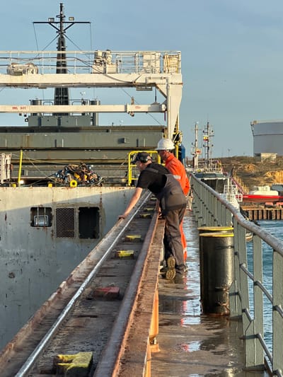 Two men are on a pier next to a large ship. One man, wearing a black t-shirt and grey overalls, is leaning over a metal railing. The second man, wearing an orange jumpsuit and a white hard hat, stands behind him, looking at the ship. The pier is wet and reflects the sky and the ship. In the background, there are other boats and industrial structures. The image appears to have been taken in daylight, with a clear blue sky. The location is the port of Uribia, Colombia.