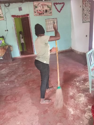 The scene is an interior room within a residence in Manda-Shivanga ward, Kenya. A barefoot individual, wearing a dark beanie, light green short-sleeved top, and dark trousers, is depicted sweeping a reddish-brown concrete floor with a long-handled broom, holding it with both hands. Dirt and swept debris are visible on the floor. In the background, a child wearing a light-colored shirt is partially visible, standing inside a brown wooden doorway. The walls are painted light blue-green and show some wear. Decorations on the left wall include a framed picture of a multi-story house with a red vehicle parked outside, a red tinsel triangle, and a smaller framed item below. A dark wooden cabinet with a patterned cloth and a television displaying a colorful image are positioned in the far-left background. A partially visible light blue-green wooden chair is on the far right. Red tinsel also adorns the top of the doorway.