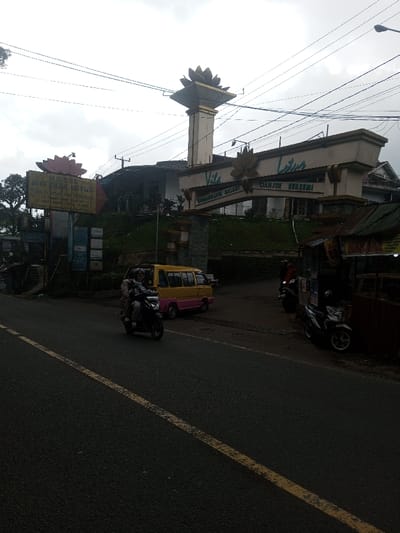 The image displays an outdoor scene, likely in Cianjur, Indonesia, based on signage. The sky is overcast. In the foreground, an asphalt road with a solid yellow line is visible.
On the left, a prominent yellow sign with a pink lotus logo advertises "HOTEL & VILLA LOTUS," displaying partially visible contact information. Behind this sign and elevated ground, a multi-story building is partially obscured by dense green foliage.
At the center, a large archway structure marks an entrance. It features a tall central column topped with a lotus flower motif, and the arch itself is adorned with "Vila Lotus," "CINDANGSARI BALADI," and "CIANJUR BERSEMI" text. Another building is visible beyond this archway.
To the right of the archway's entrance, a yellow and pink minivan is situated, either parked or in motion within the driveway area. Further right, a small stall with a corrugated metal roof and wooden frame stands beside the road, displaying various items and posters. Two scooters are parked adjacent to this stall.
On the road in the foreground, a dark-colored scooter is actively moving, carrying two individuals, both wearing helmets. Utility lines crisscross the sky above the entire scene.