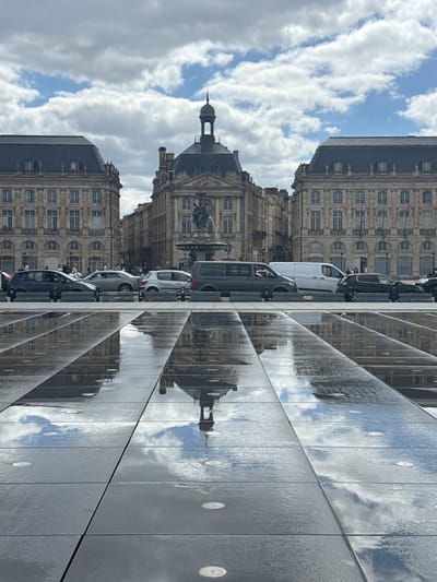 The image depicts the Place de la Bourse in Bordeaux, France, a historic public square. In the foreground, a reflective pool of water is laid out on large, dark tiles, creating mirror images of the sky and the surrounding architecture. The sky is partly cloudy, with patches of blue peeking through.

Behind the reflective pool, a row of cars is parked, including a dark hatchback, a silver sedan, a white van, and a dark minivan. Some people are visible in and around the cars and walking in the background.

The background features the iconic architecture of the Place de la Bourse. Two grand, stone buildings with mansard roofs and numerous windows flank a central building. The central building is characterized by a dome-shaped roof topped with a spire and a statue at its base, a fountain. The buildings exhibit classical architectural details. The overall scene is bright and airy, suggesting it was taken on a clear day.