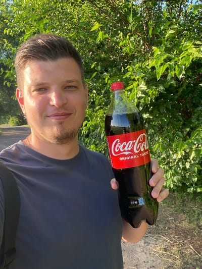 In Yablunivka, Ukraine, a young man stands outdoors in front of a lush green tree line, holding a large, two-liter bottle of Coca-Cola. The man, with light skin and short, dark hair, is wearing a dark blue t-shirt and a gold chain. He is smiling and looking towards the camera, his right hand gripping the bottle. The Coca-Cola bottle is partially filled with a dark liquid and has its distinctive red label with white lettering. The background consists of dense foliage and a hint of a dirt path and road to the left, suggesting a rural or semi-rural setting. The lighting indicates it is daytime, with sunlight casting shadows and highlighting the man's features and the bottle.