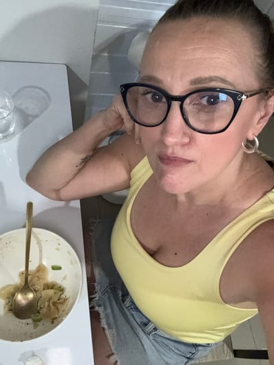 A woman with dark hair pulled back, wearing black-rimmed glasses, a yellow tank top, and jean shorts, poses for a selfie. She is seated at a white table with a partially eaten bowl of pasta, a gold spoon, and a glass of water. The location in Moo 4, Thailand is not discernable from the image.