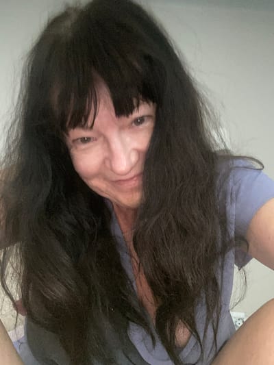 A selfie of a woman with long, dark, wavy hair and bangs. She is wearing a light blue t-shirt and looking at the camera with a slight smile. The background is plain and light-colored, suggesting an indoor setting. The image does not contain information about the city of Novi Petrivtsi, Ukraine.