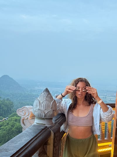 In Tây Hoa Lư Ward, Vietnam, a woman with curly brown hair stands on a balcony overlooking a misty valley. She is wearing a light pink crop top, an open light blue shirt, and olive green pants. She is holding a pair of sunglasses up to her eyes. The balcony railing is made of stone and features intricate carvings, including a lotus flower design. In the background, a mountain peak emerges from the fog, and lush green trees cover the slopes. The sky is overcast and a pale blue.