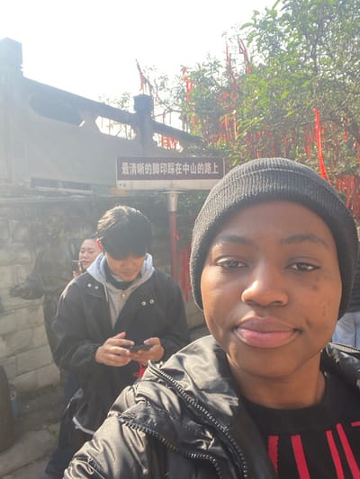 A person wearing a beanie is taking a selfie outdoors. Behind them is another person looking at a phone, and a third person is partially visible. There are trees with red ribbons tied to them and a sign with Chinese characters. The area appears to be a park or garden on a sunny day. The picture is taken from a low angle, giving a perspective of looking up at the subject.