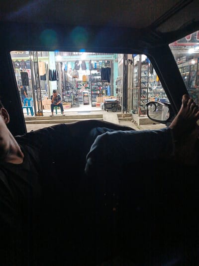 The image shows a view from inside a vehicle, looking out at a street scene at night in Tingkeummanyang, Indonesia. In the foreground, a person's legs are visible, resting on the dashboard or seat of the car. The person appears to be relaxed, with their feet propped up.
Outside the car, a street is lined with shops. The shops are illuminated, and displays of clothing and merchandise are visible through open doorways and windows. One shop appears to be selling apparel, with racks of shirts and pants on display. Another shop seems to be a general store or department store, with shelves stocked with various items, including what look like shoes and bags. A few people are visible in front of the shops. One man is sitting on a blue chair, looking down. Another person is standing near a display of bags. A blue suitcase is also visible.
The overall impression is of a busy street scene at night, with commerce in full swing. The lighting suggests that it is likely evening or nighttime. The presence of shops and people indicates an urban or semi-urban environment. The specific location cues are consistent with the provided context of Tingkeummanyang, Indonesia. The mood seems casual and relaxed, given the posture of the person in the foreground.