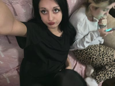 This image depicts an indoor scene in Lanchkhuti, Georgia, featuring two females. In the immediate foreground, a female with long dark hair, dark eyes, and red lipstick is visible, facing the camera with a pursed lip expression. She is wearing a black short-sleeved top, and her right arm extends partially into the frame, suggesting she is holding the camera device. To her right and slightly behind, a second female with blonde hair is seated, looking down at a smartphone with a glowing screen. She is wearing a light grey long-sleeved top and leopard-print patterned pants. White socks are visible on her feet. Both individuals are situated on what appears to be a bed or sofa; the surface beneath the foreground female is a light pink fabric with a faint tree branch pattern, while the second female rests on the leopard-print fabric and a section of the pink patterned material. The overall environment suggests a casual, relaxed setting.